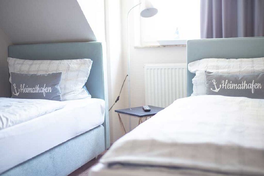 Ein Hotelzimmer mit zwei Betten, jeweils mit Kopfstütze und Kissen. Es gibt einen kleinen Tisch mit Lampe und Fernbedienung.