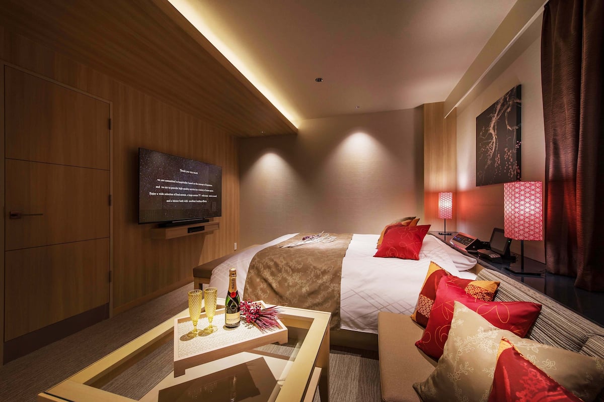 Een hotelkamer met een bed, een televisie, een bureau met een fles en glazen, en een rode lamp.