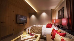 Une chambre d’hôtel avec un lit, un téléviseur fixé au mur, une table de chevet avec une bouteille et des verres, et un canapé aux motifs rouges et dorés.