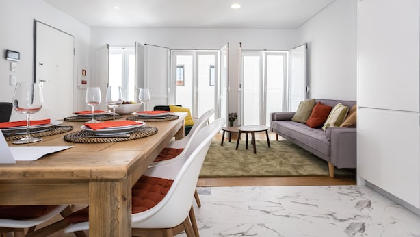 A modern living room with a wooden dining table, white chairs, and a grey sofa.