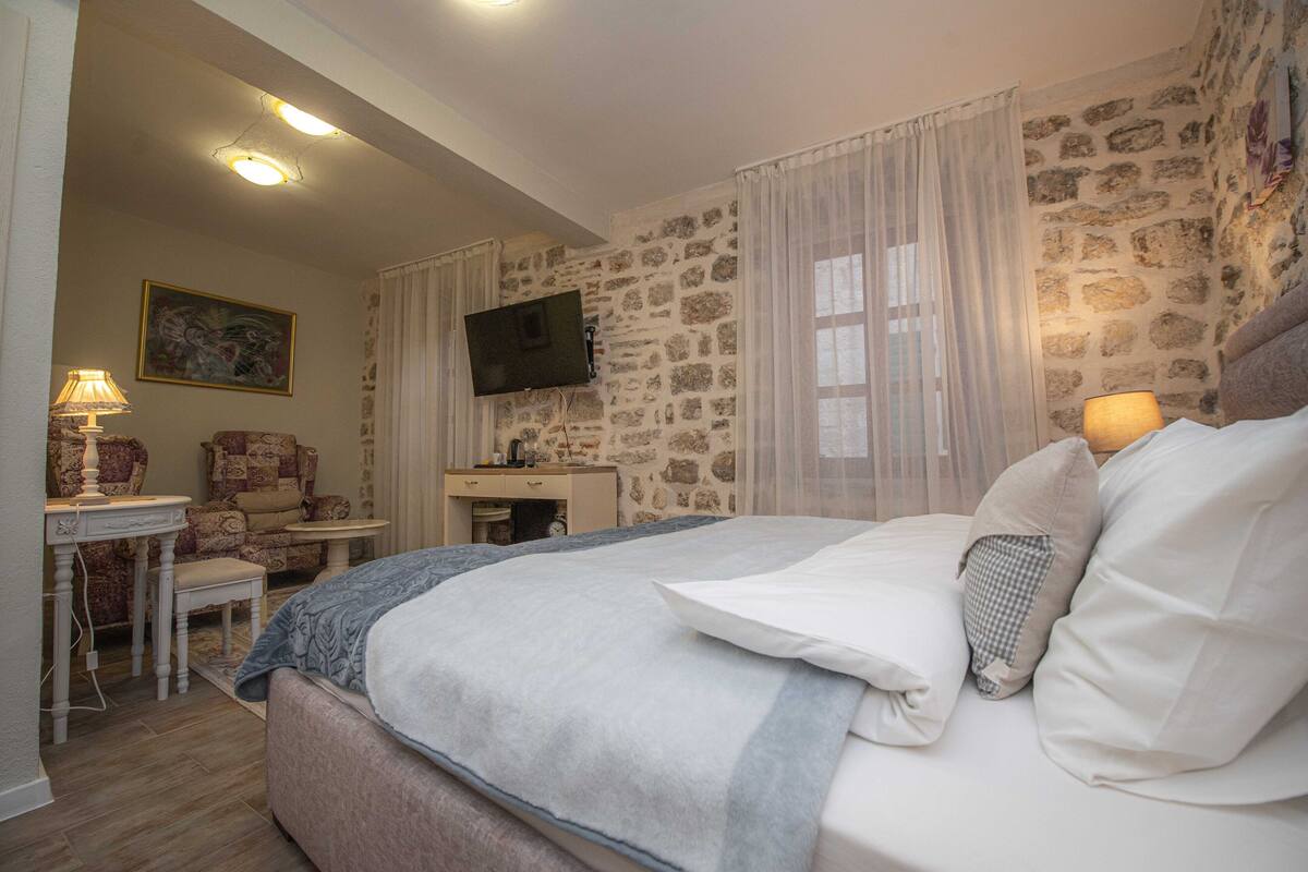 Quarto com parede de pedra, cama com roupa de cama branca, televisão na parede e escrivaninha com luminária.