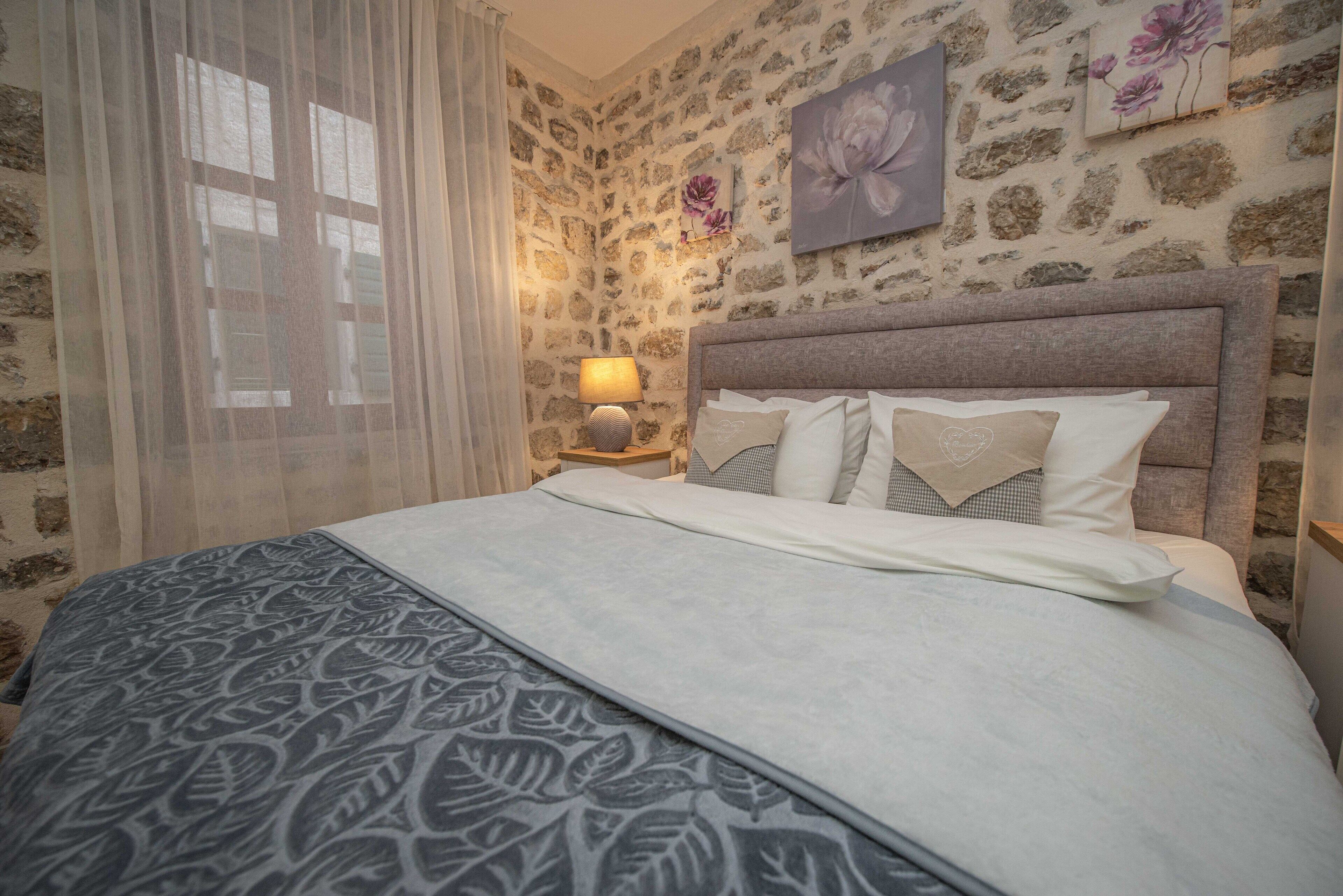 Ein Schlafzimmer mit Steinwand, einem Bett mit gemusterten Betttextilien, einer Nachttischlampe und floralen Kunstwerken.