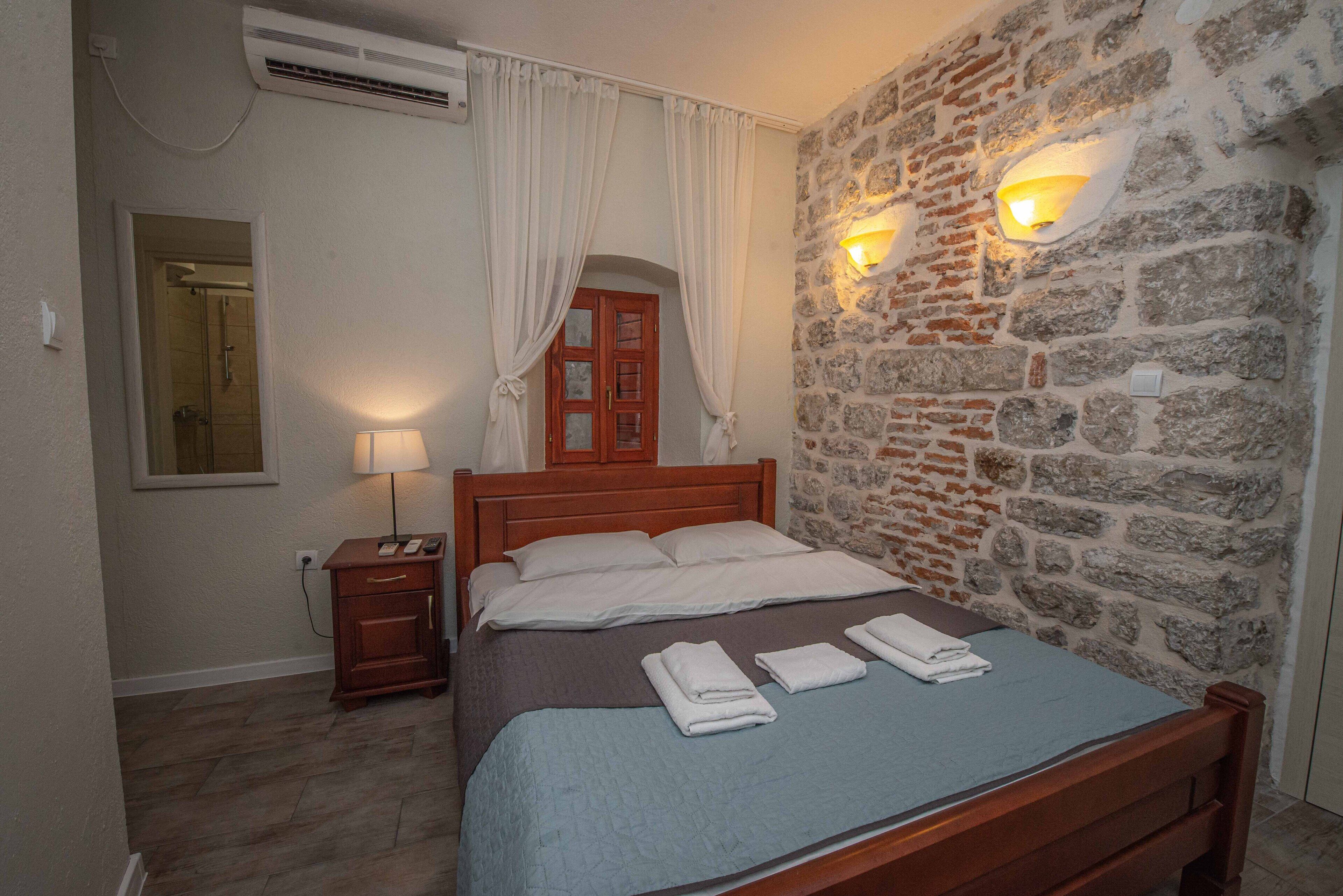 A bedroom with a stone wall, a wooden bed, a nightstand, a lamp, and a mirror.
