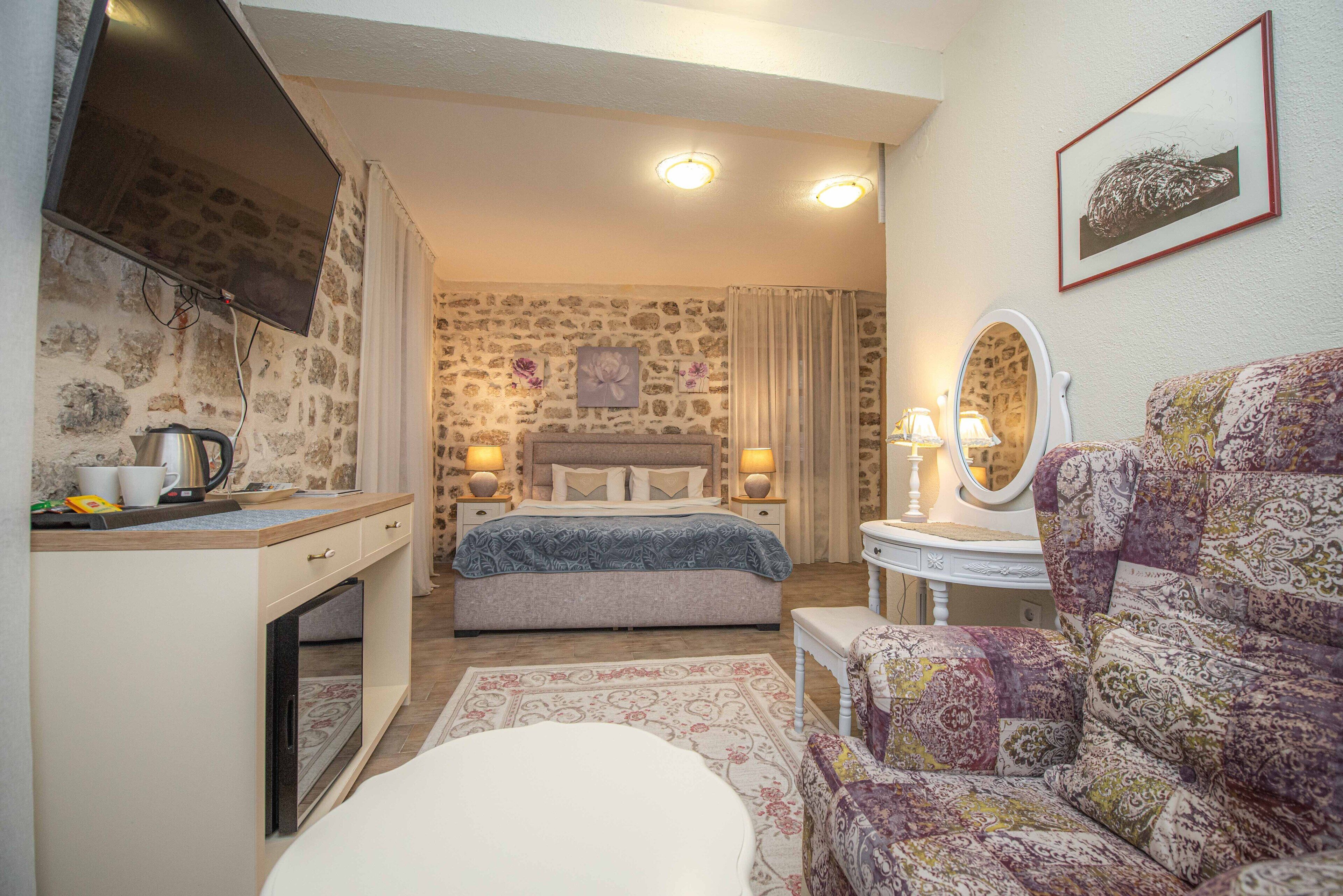 A bedroom with a stone wall, a bed with a blue blanket, a bedside table with a lamp, a patterned armchair, and a small table with a mirror.
