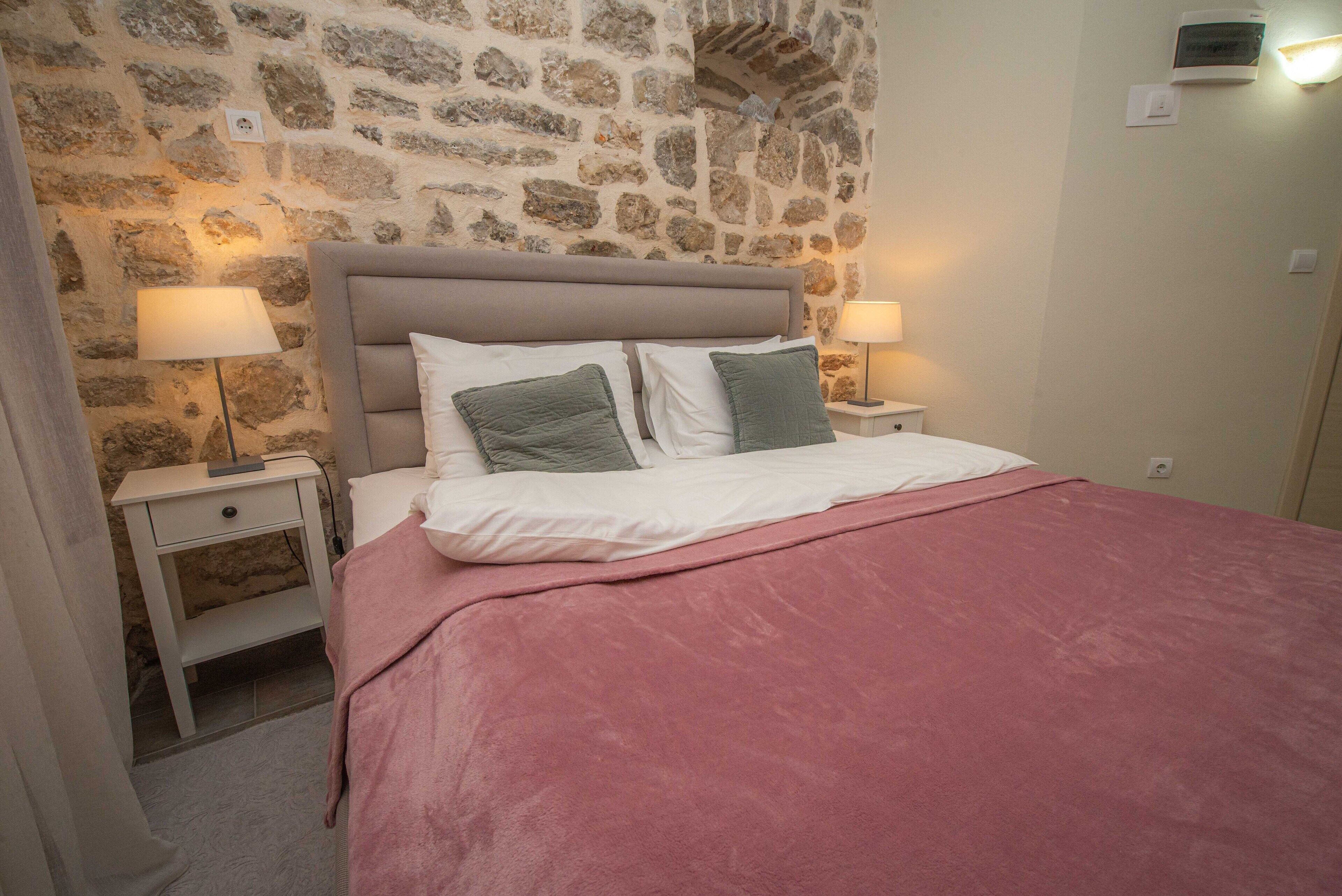 A bedroom with a stone wall, a bed with a pink blanket, two bedside tables with lamps, and a nightstand with a drawer.