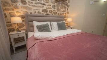 A bedroom with a stone wall, a bed with a pink blanket, two bedside tables with lamps, and a nightstand with a drawer.