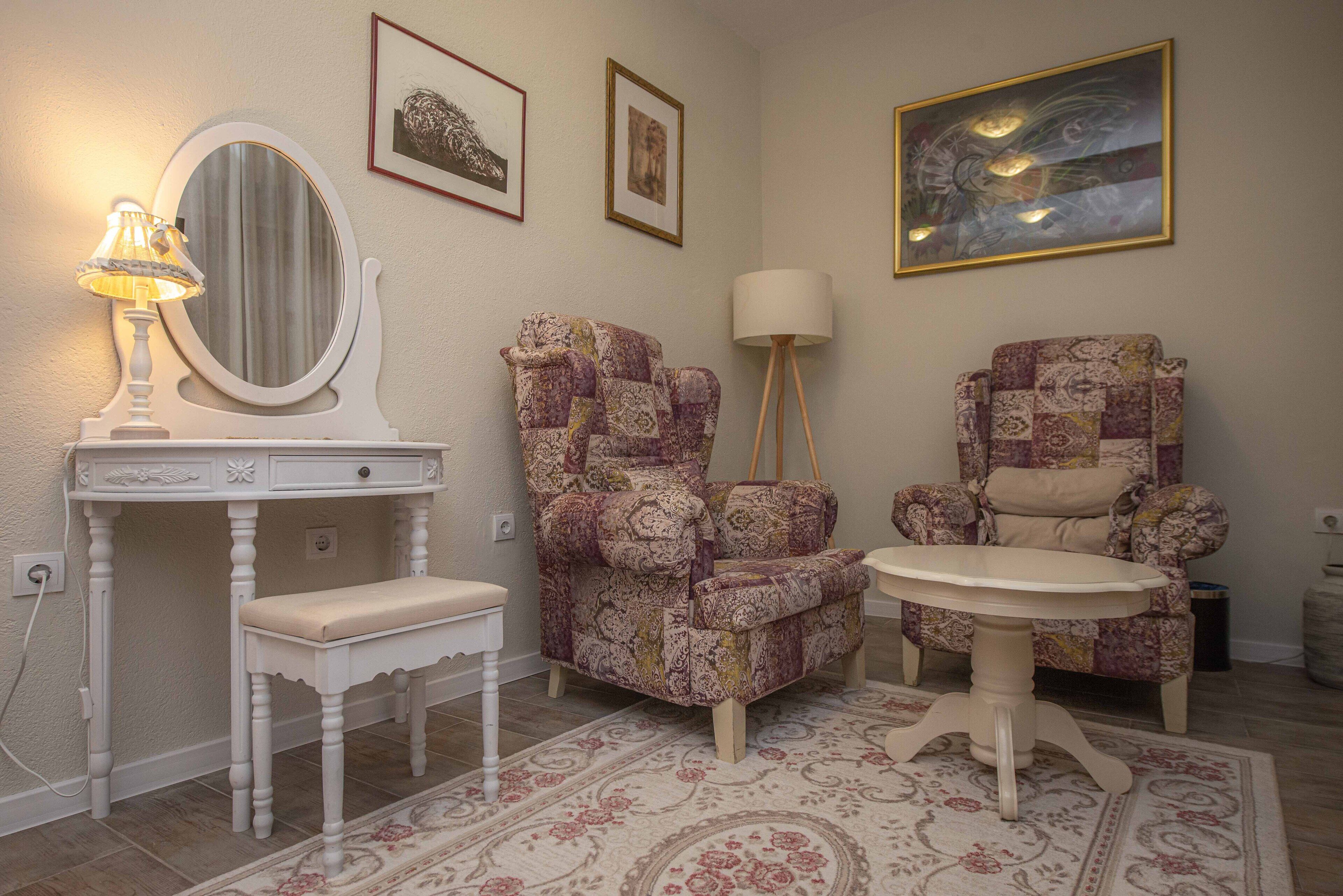 A room with a white vanity, two patterned armchairs, a small round table, and a large framed painting on the wall.