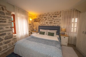 Un dormitorio con pared de piedra, cama con cubrecama azul, almohadas blancas, mesita de noche con lámpara y armario.