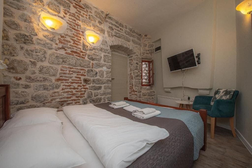 A room with a stone wall, two beds with white linens, a TV mounted on the wall, and a small table with a chair.