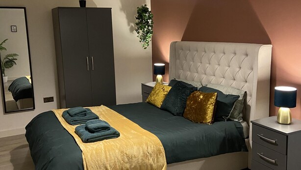 A modern bedroom with a tufted headboard, a large bed with dark green and yellow bedding, a grey wardrobe, a hanging plant, and a bedside table with a lamp. - Captivating 2-bed Apartment in Bradford (Bradford)