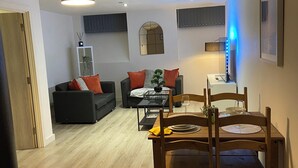A modern living room with a dining area, featuring a wooden dining table with chairs, a sofa with orange cushions, a small side table, and a ceiling light fixture. - Captivating 2-bed Apartment in Bradford (Bradford)