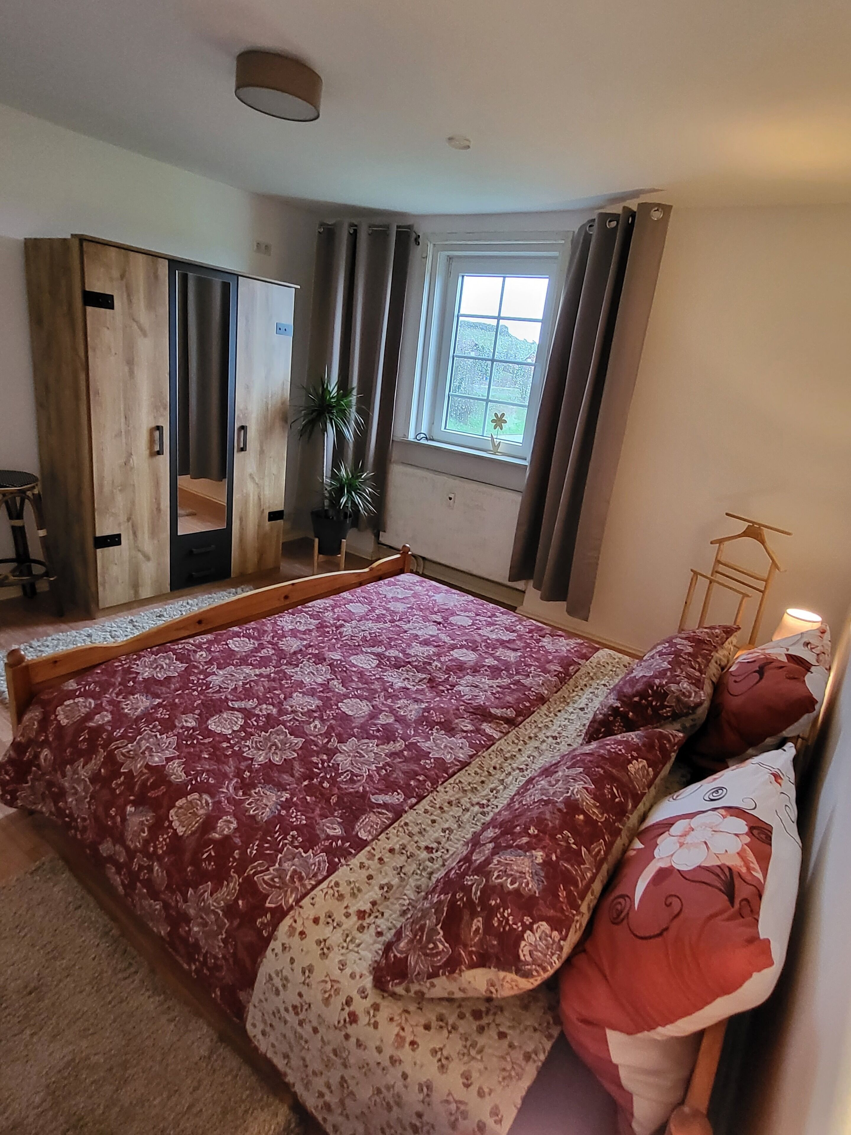 Een slaapkamer met een groot bed, een kledingkast, een klein tafeltje en een raam met gordijnen.