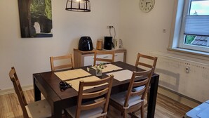A dining area with a wooden table, four chairs, a coffee maker, a clock, and a window with blinds.