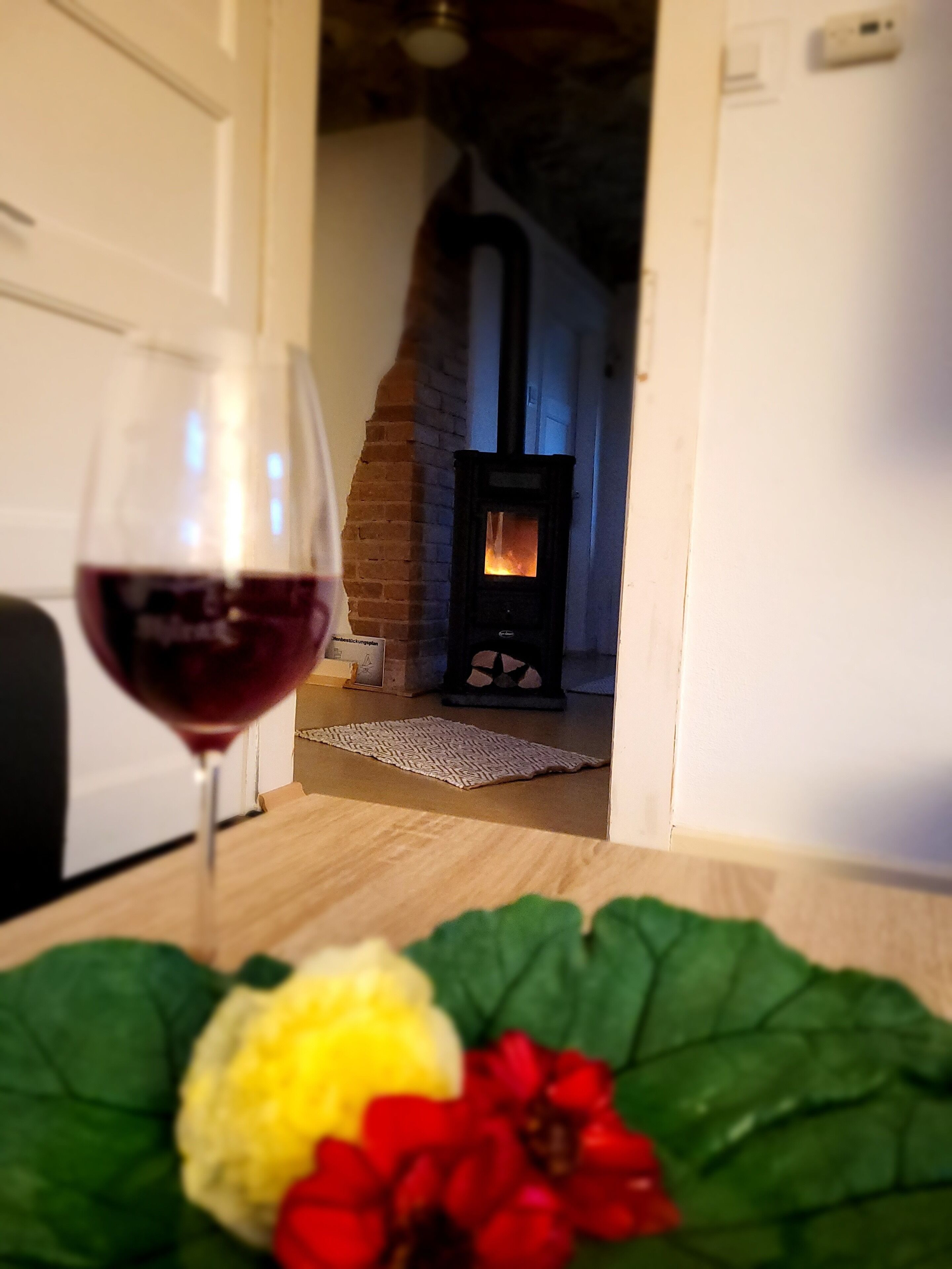 A room with a fireplace, a glass of red wine, and a table with flowers.