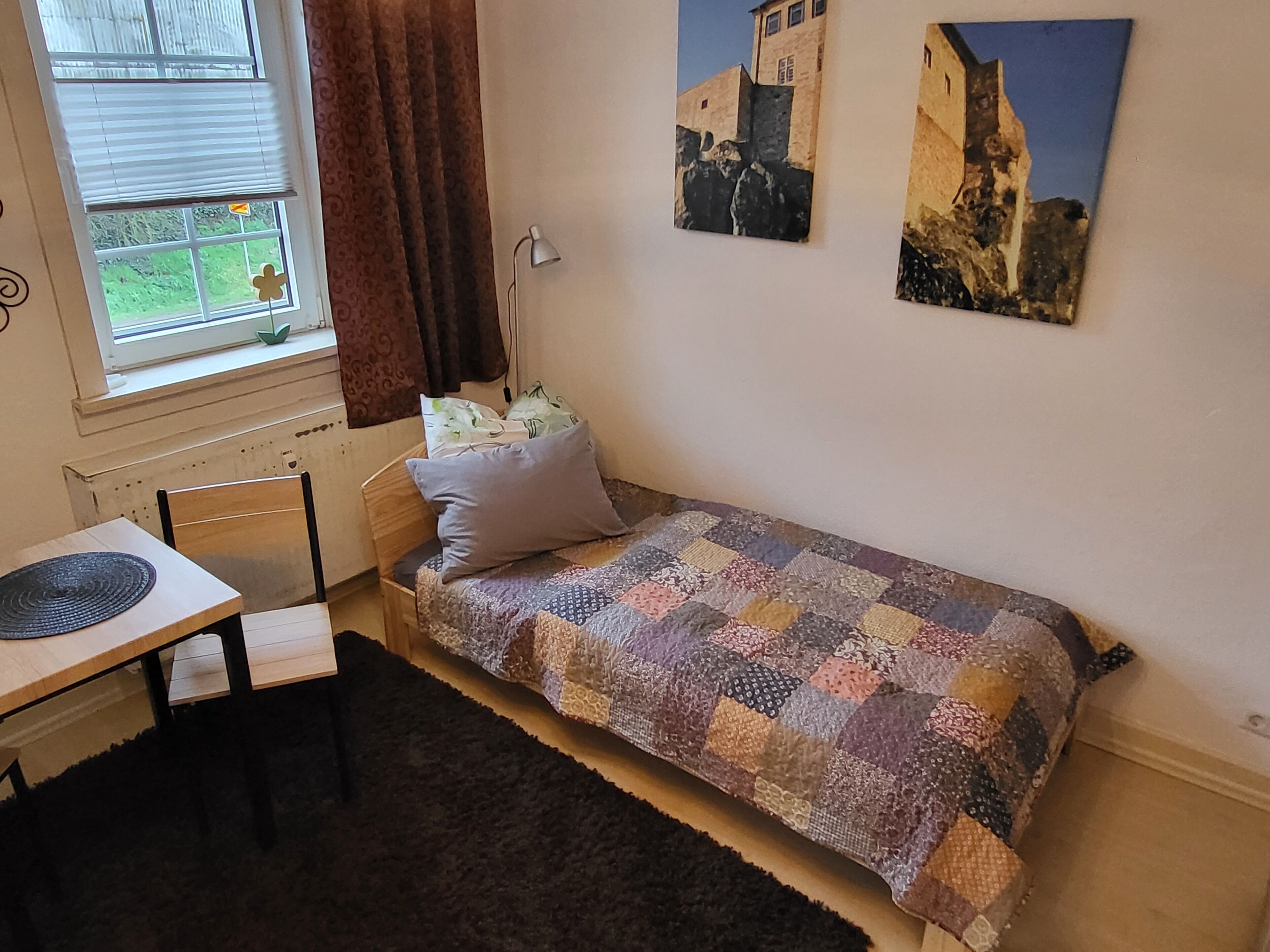 Une petite chambre avec un lit simple, un bureau et une chaise. Il y a deux tableaux au mur, une fenêtre avec des rideaux et un radiateur.