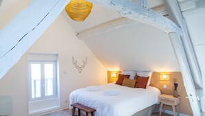 Ein Schlafzimmer mit schrägem Dach, einem Bett mit weißer Bettwäsche, einer Holzbank, einem kleinen Tisch mit einer Vase und einem Fenster mit Jalousien.