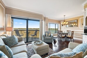 A spacious living room with a large sliding glass door leading to a balcony, a dining area with a chandelier, and a kitchen with a large mirror. - Spectacular Villa on Broad Creek w/ Pool, Hot Tub Walk to Shops at Shelter Cove! (Hilton Head Island)
