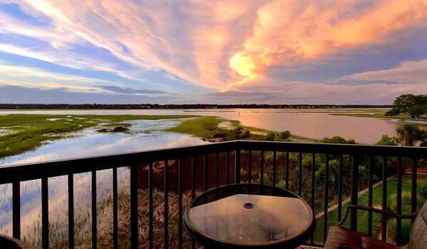 A deck with a glass-top table and a view of a river and fields.