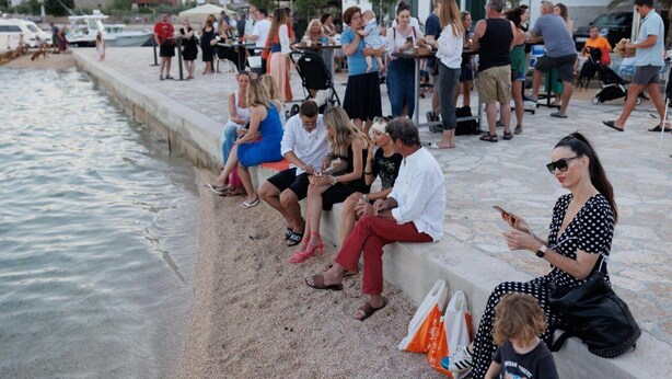 A group of people sitting by a waterfront promenade with a view of the sea and buildings in the background.