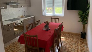 A compact kitchen with a modern stove, a sink, and wooden cabinets. There is a dining table with a red tablecloth and four wooden chairs. A window with blinds is visible, and a potted plant is placed near the window.