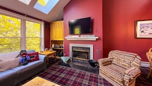 A living room with a fireplace, a sofa, a television mounted above it, and a window with a view of trees. - Cozy 2 BR Ski-in/Ski-out Condo at Okemo (Ludlow)