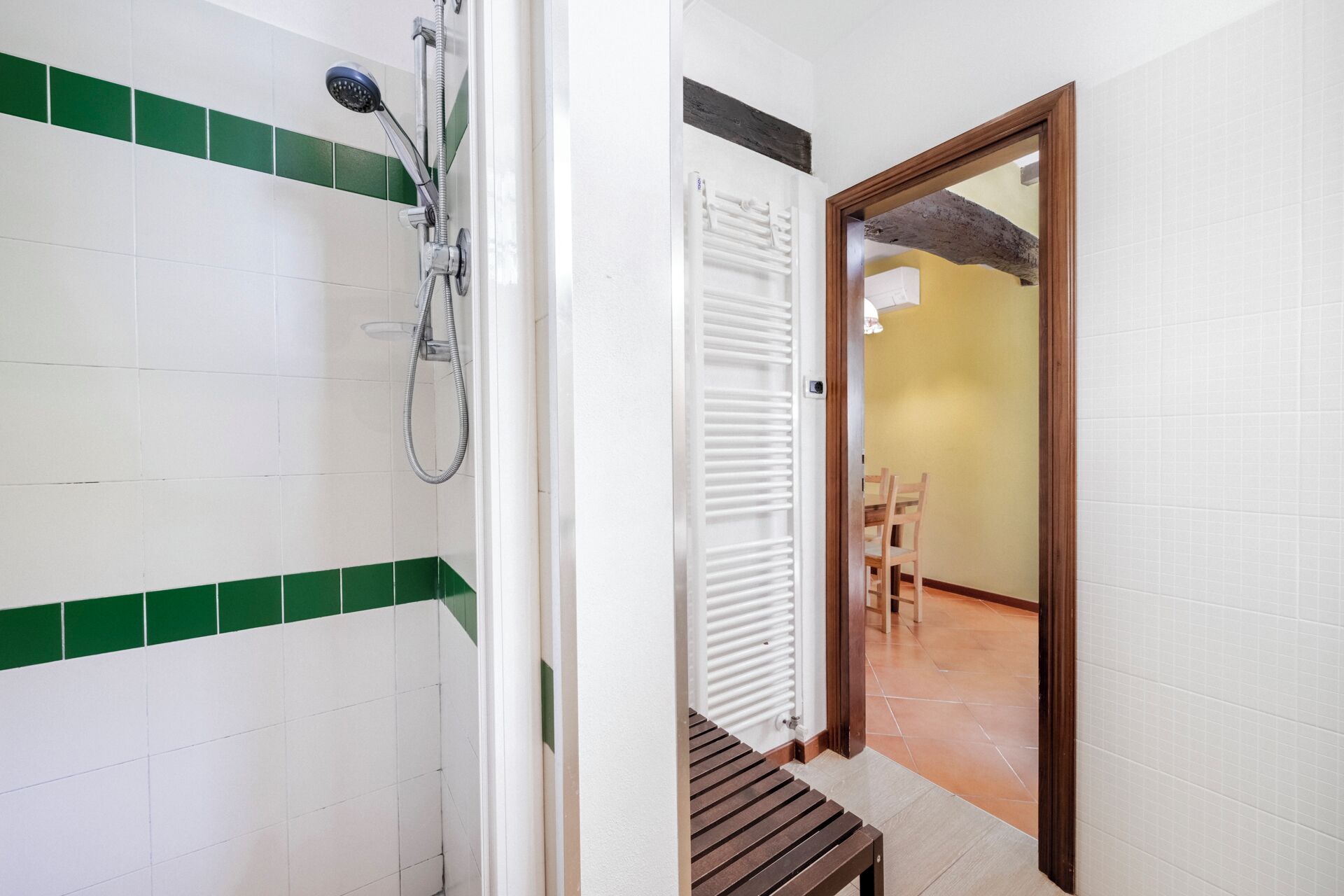 Un baño con azulejos blancos y verdes, una ducha, un radiador blanco y un banco de madera.