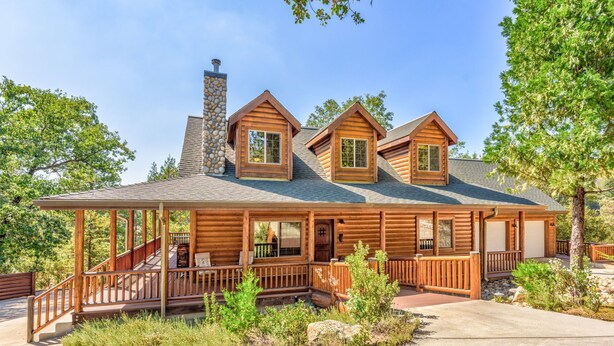 A log cabin with a covered porch, a stone chimney, and a landscaped front yard.