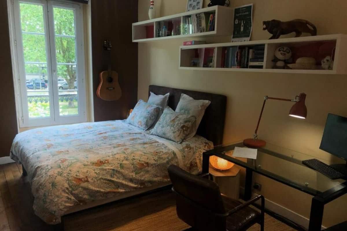 A bedroom with a bed, a desk with a computer, a chair, a guitar, and shelves with books and decorative items.