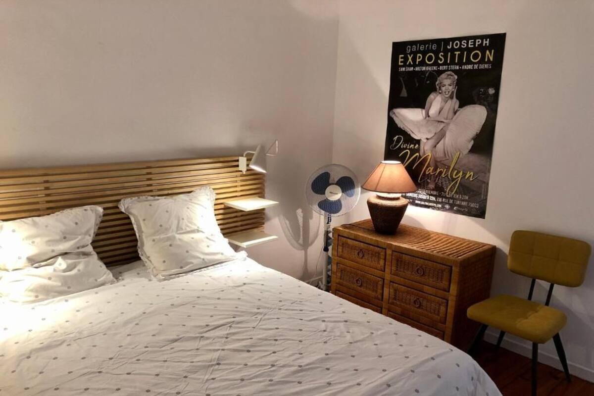 A neatly made bed with a patterned bedspread, a wooden headboard, a bedside table with a lamp, a fan, and a poster on the wall.
