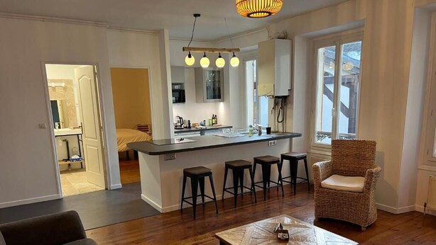 A modern kitchen with a central island, bar stools, and a wicker chair. - Villa Eugénie, Gîtes & Chambres d'Hôtes  (Melun)