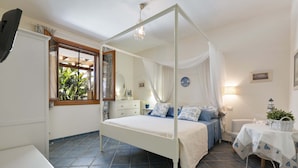 Un dormitorio con una cama con dosel, una mesita de noche, una lámpara de noche, un ventanal con vistas a la vegetación y un reloj de pared.