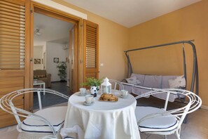 Une table ronde recouverte d’une nappe blanche, quatre chaises blanches, un plateau de pâtisseries, deux tasses blanches et une petite lanterne blanche.