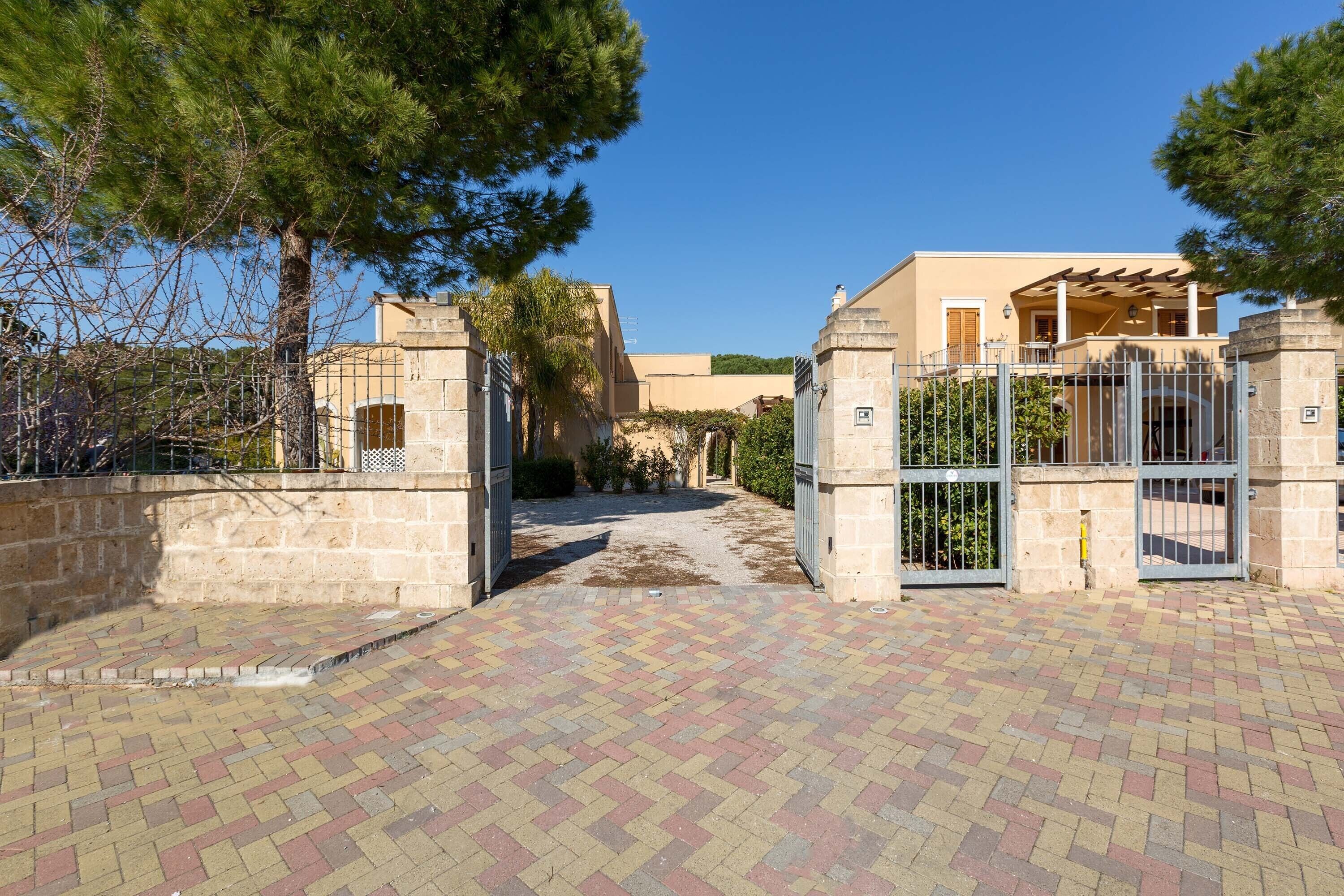 A paved entrance with metal gates, a stone wall, and a building with a beige facade and a covered porch.