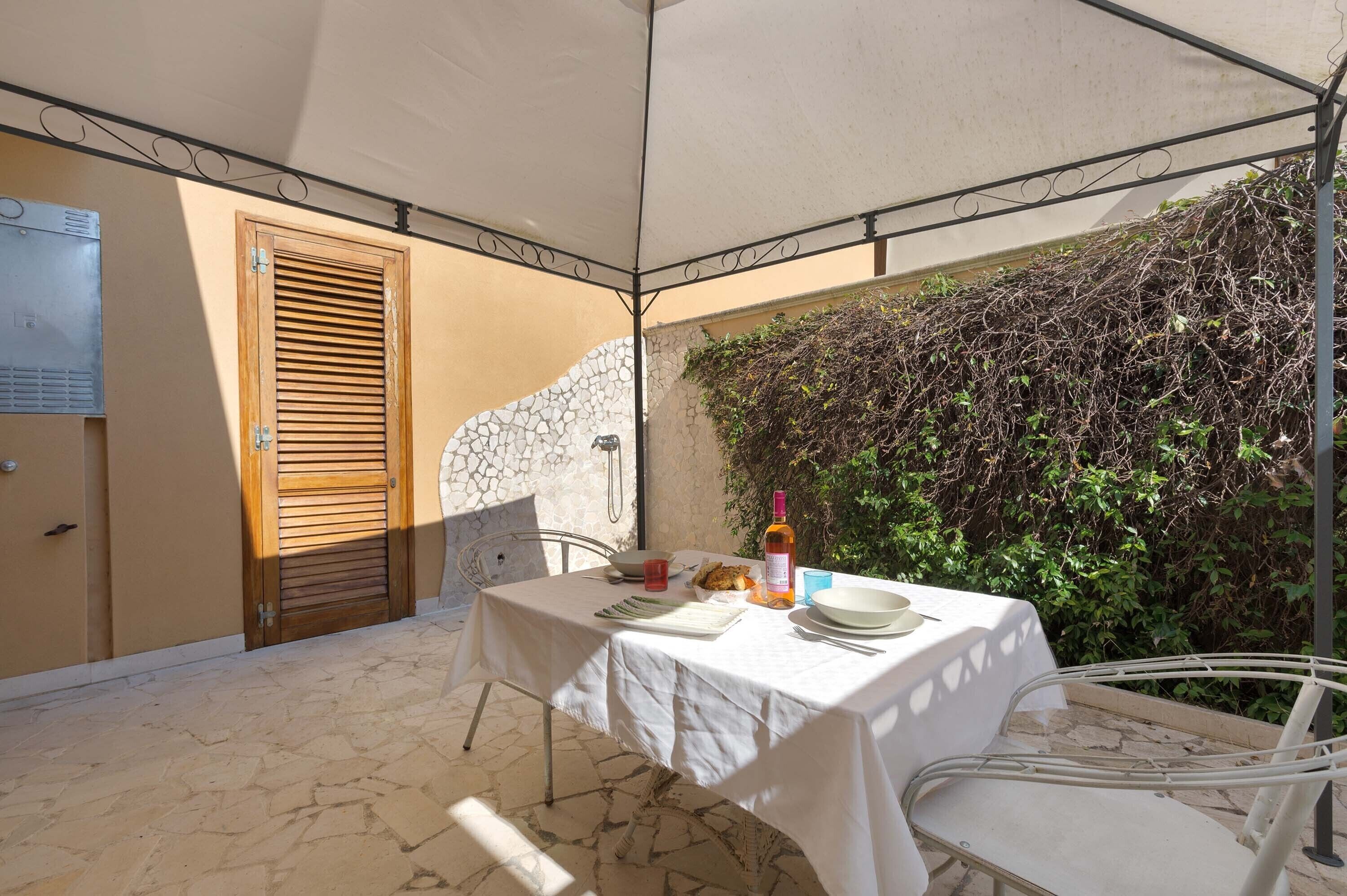 Un espace extérieur abrité, aménagé pour prendre les repas, avec une table dressée pour deux personnes, une bouteille de vin et une assiette de nourriture.