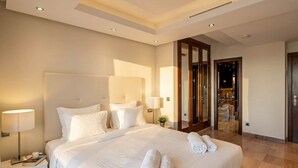 A modern hotel room with a large bed, a nightstand, a lamp, and a view of the hotel's interior through a glass door.