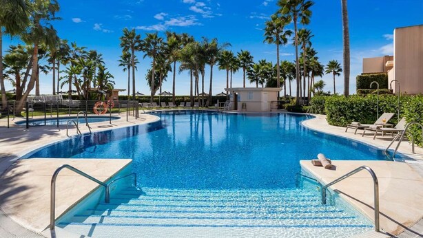 A swimming pool surrounded by palm trees and lounge chairs, with a clear blue sky above.