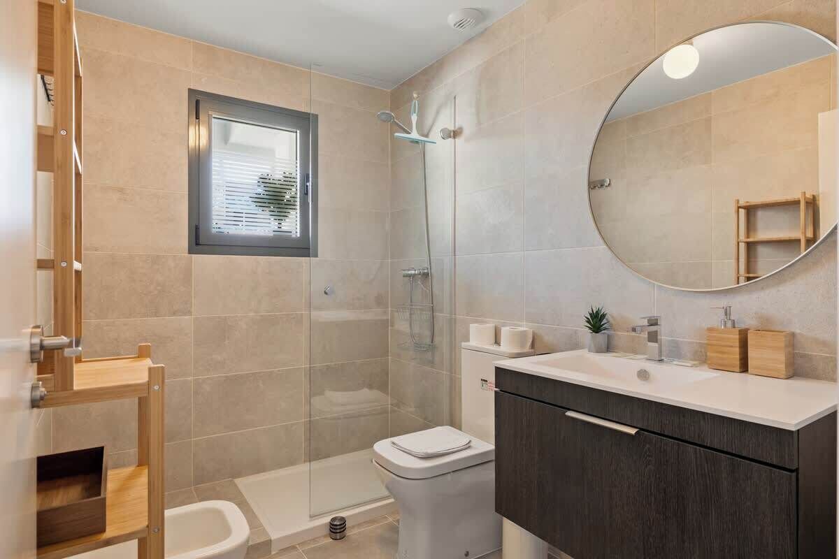 A modern bathroom with a glass-enclosed shower, a toilet, a sink with a mirror, and a window with blinds.