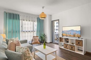 Un salon moderne avec un canapé, une table basse, un téléviseur posé sur un meuble blanc et une vue sur un immeuble par la fenêtre.