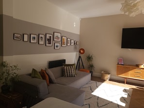 A modern living room with a grey sectional sofa, a mounted flat-screen TV, and a wall adorned with framed pictures. - Beautiful Family Home in the heart of Sheffield (Sheffield)