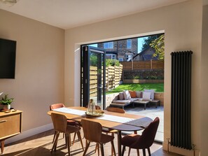 A modern dining area with a large table and chairs, a view of a backyard with a patio and outdoor seating, and a sliding glass door leading to the outside. - Beautiful Family Home in the heart of Sheffield (Sheffield)