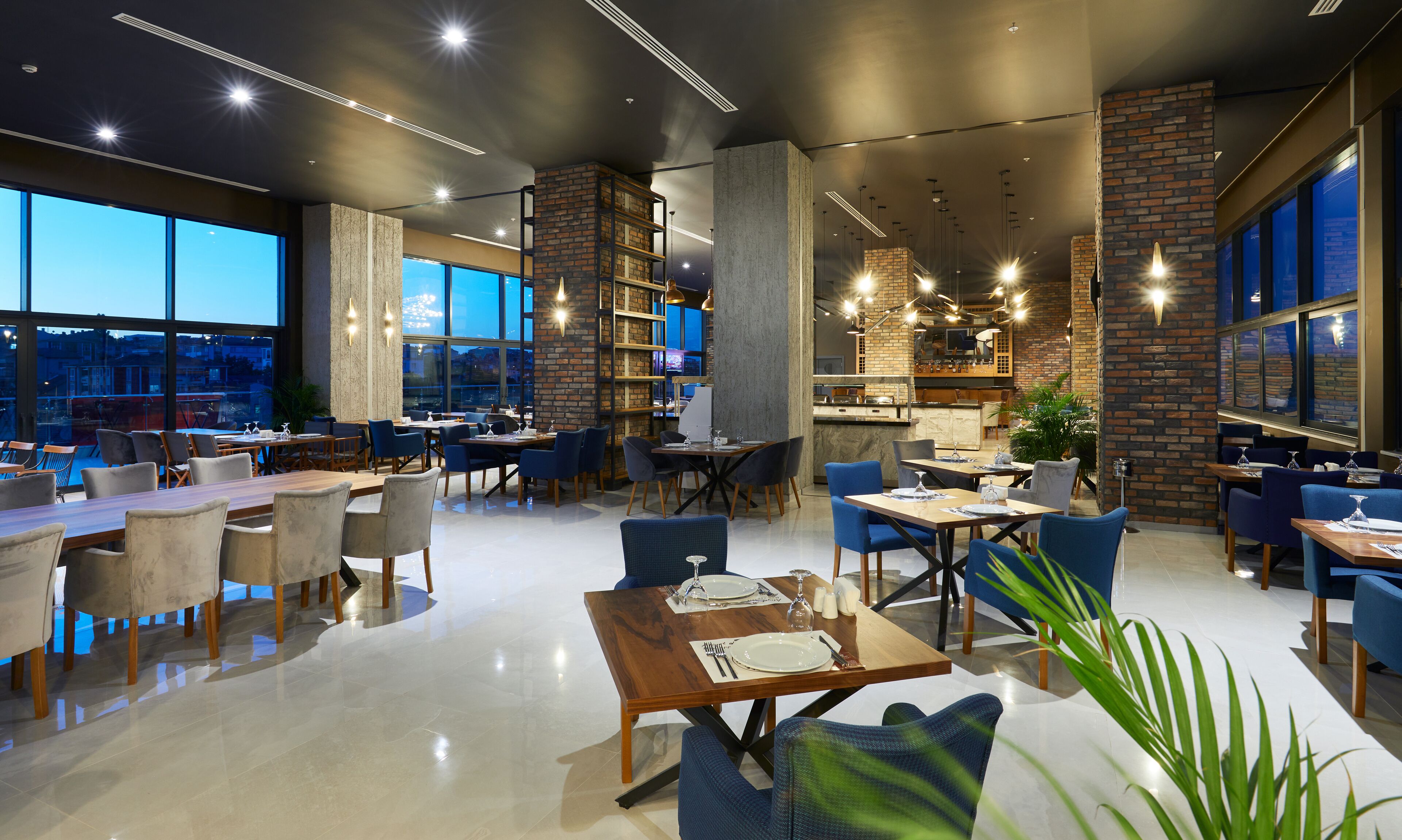 A modern restaurant with high ceilings, exposed brick walls, and large windows offering city views. Tables are set with white tablecloths and blue chairs.