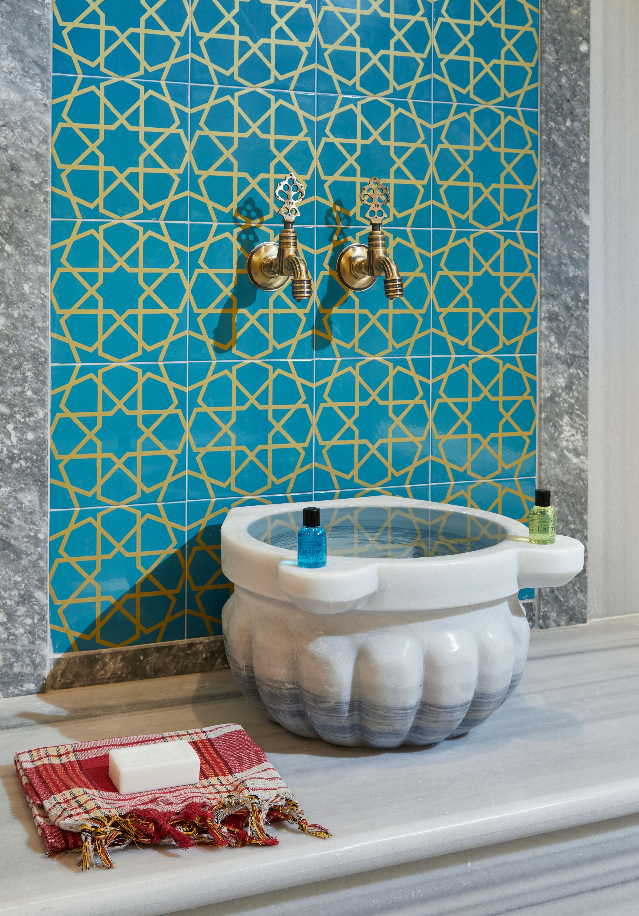 A bathroom with a blue and gold patterned tile wall, a white marble sink, and a red and white towel.