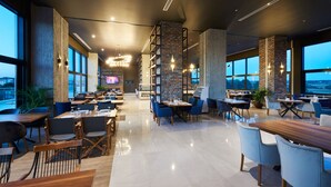 Ein modernes Restaurant mit hohen Decken, freiliegenden Backsteinwänden und großen Fenstern mit Blick nach außen.