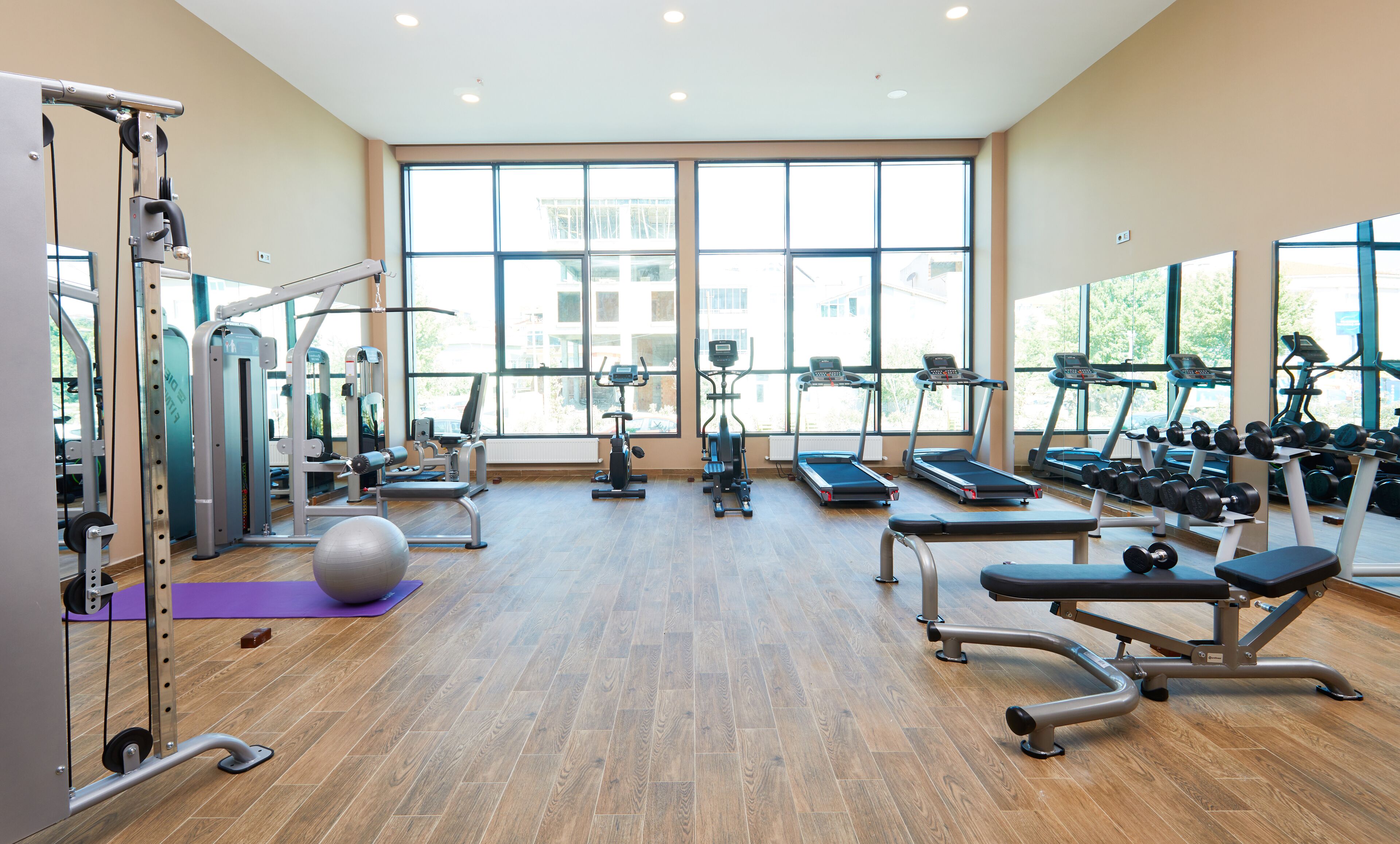 Ein großzügiger Fitnessraum mit unterschiedlichen Trainingsgeräten, großen Fenstern und Holzboden.