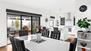 A modern kitchen with white cabinetry, a central island, and a dining area with a white table and black chairs. - Muirfield Beach House 3a (Jan Juc)