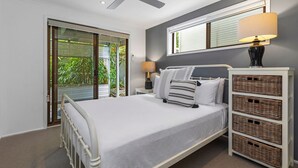 Een slaapkamer met een bed, nachtkastje, lamp, raam met jaloezieën en uitzicht op groen.