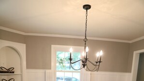 A dining room with a wooden table set for six, white chairs, a chandelier, and a window with blinds.