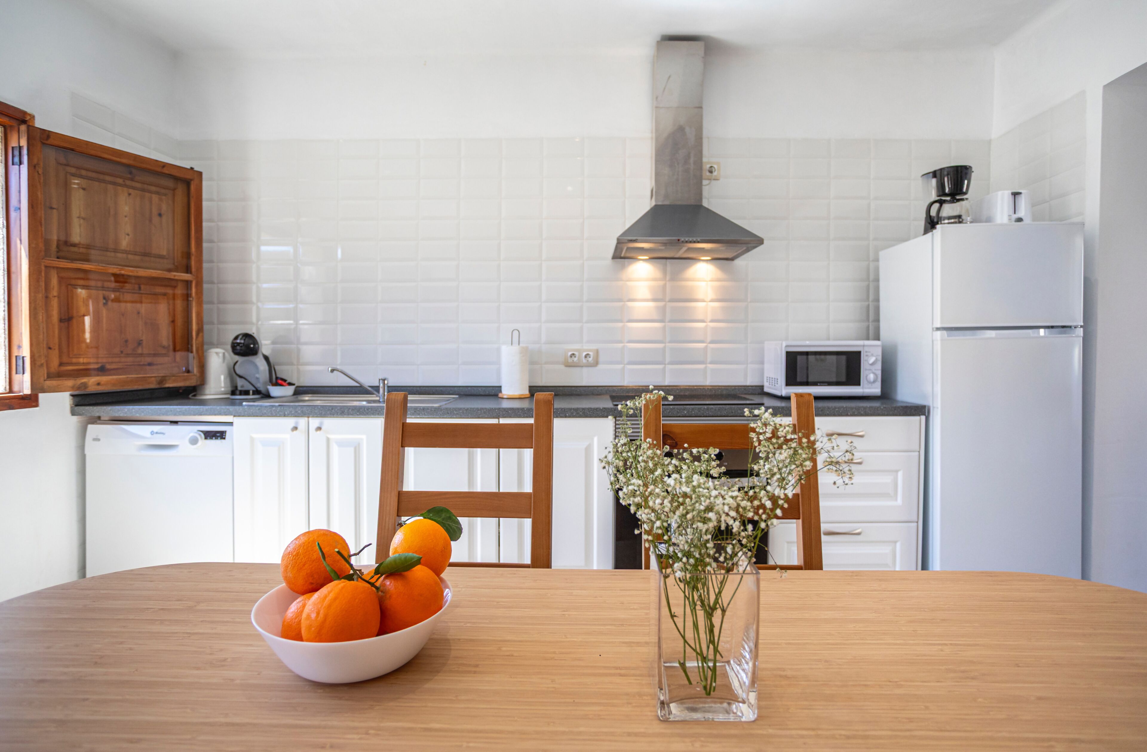 Una cocina moderna con armarios blancos, un microondas, una estufa y una mesa de comedor con un bol de naranjas y un jarrón de flores.
