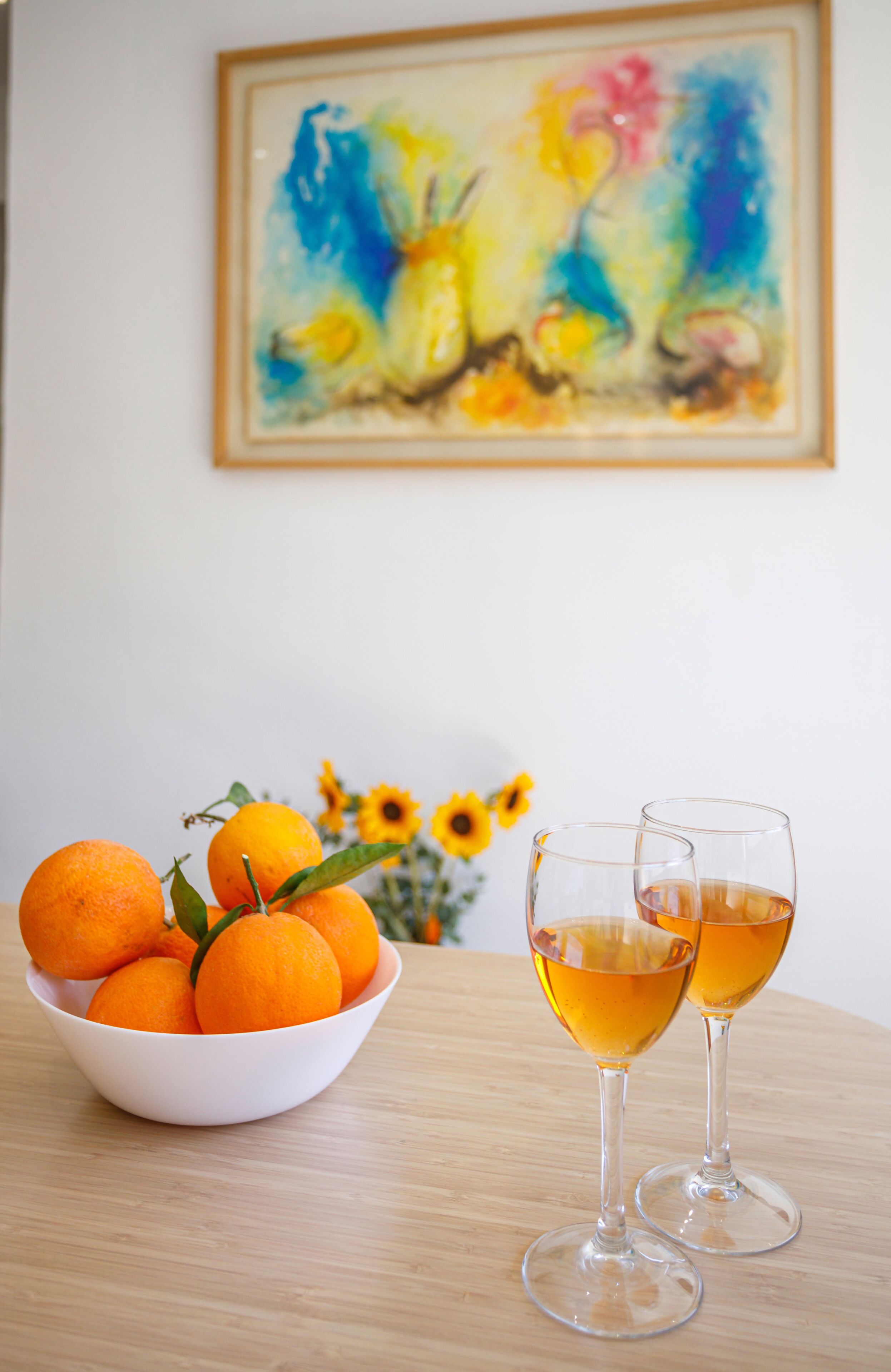 A bowl of oranges and two glasses of a yellowish beverage on a wooden table, with a painting and sunflowers in the background.