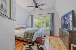 A bedroom with a bed, a ceiling fan, a TV, a window with curtains, and a wooden floor.